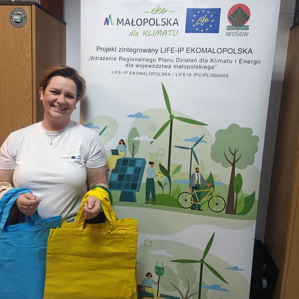 Uśmiechnięta kobieta na tle roll-upu z grafiką i logotypem projektu z bawełnianymi torbami  