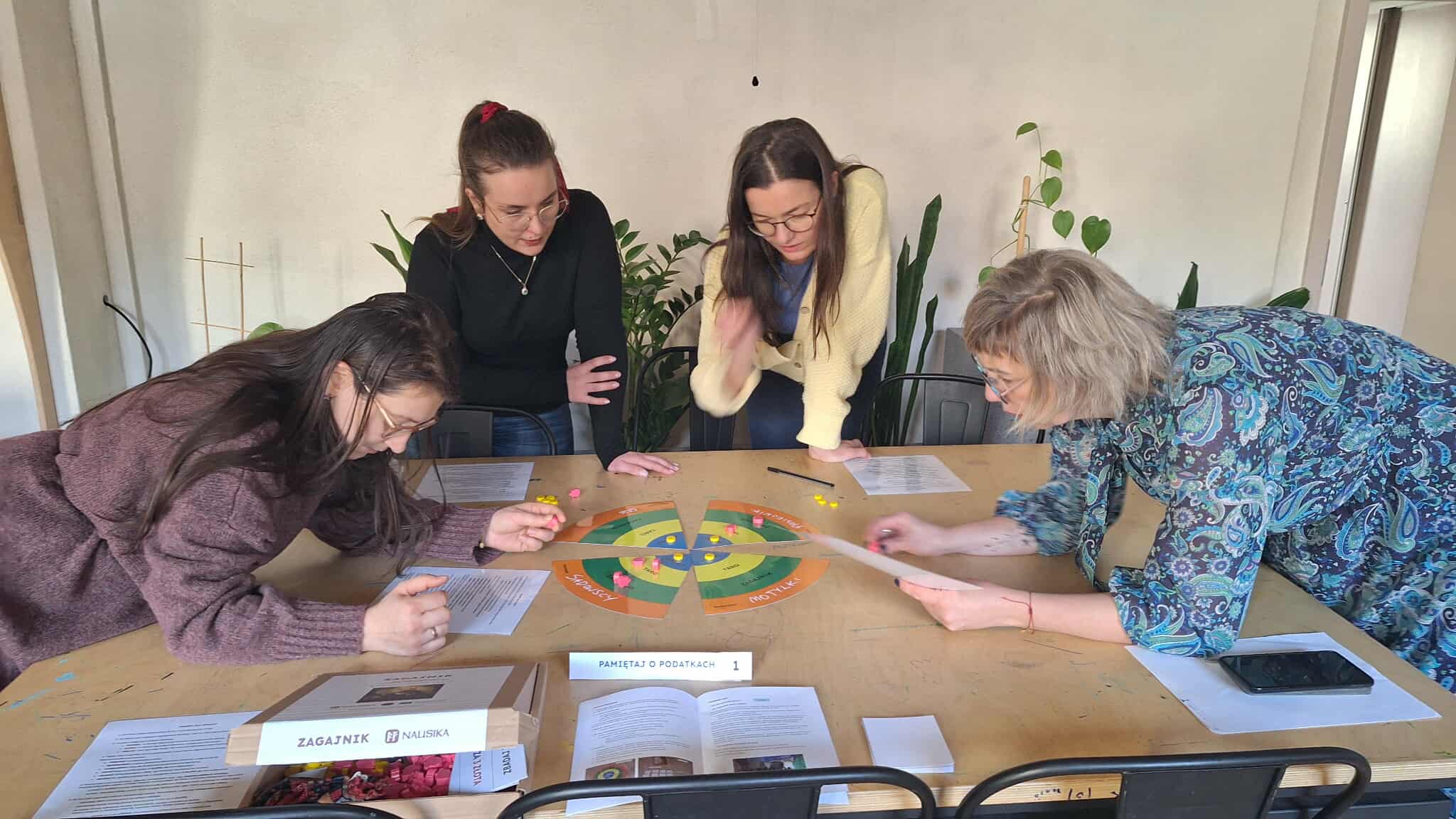 Cztery kobiety stoją pochylone nad stołem i analizują grę ekologiczną, która będzie wykorzystywana na zajęciach z dziećmi  