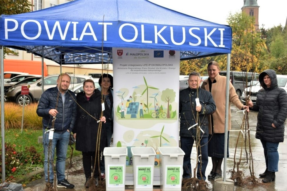 Grupa ludzi stoi na stoisku trzymając sadzonki drzew na tle roll-upu z logotypem projektu