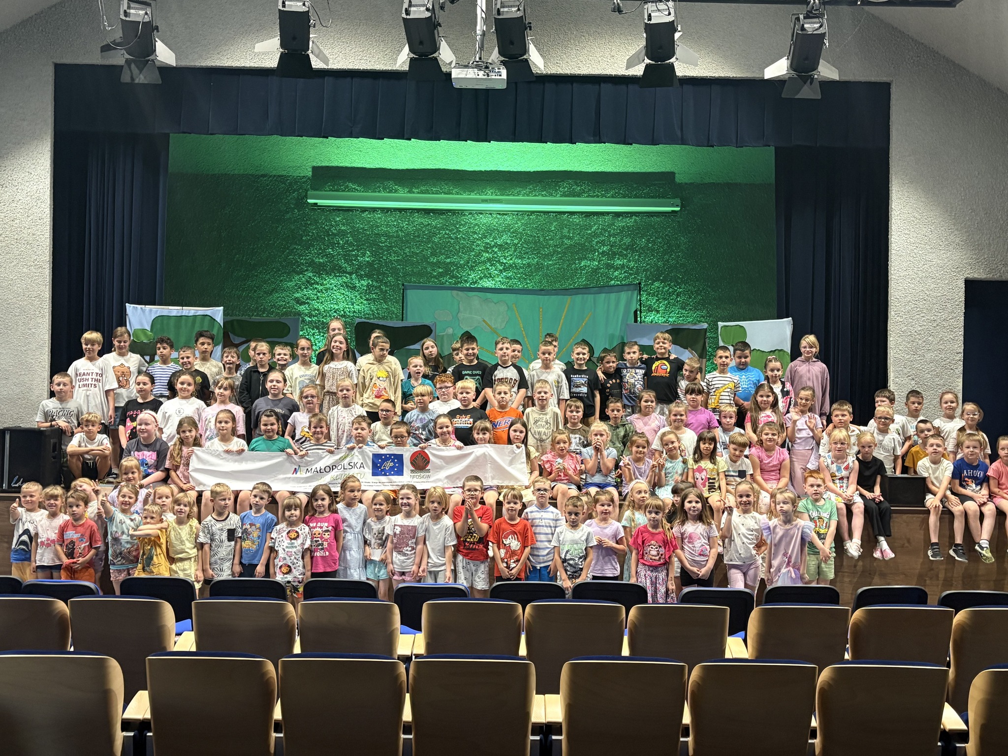 Grupa dzieci po spektaklu ekologicznym stoi na scenie trzymając baner z logotypem projektu 