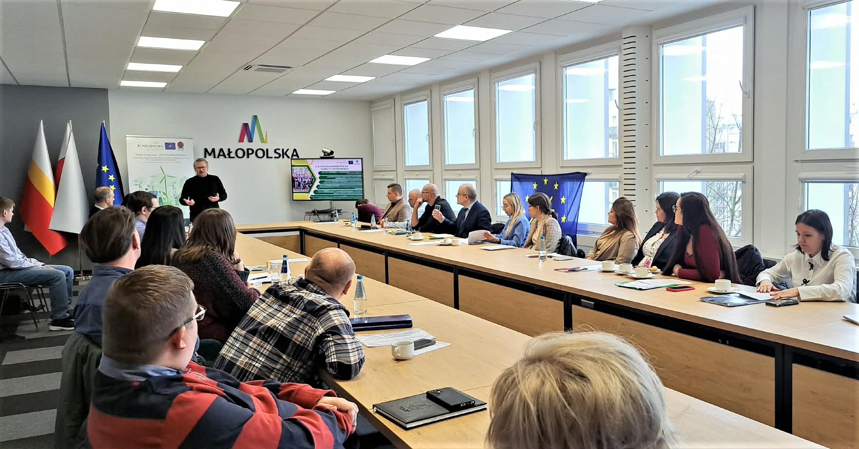 większa grupa ludzi siedzi przy stole w kształcie litery U i rozmawia