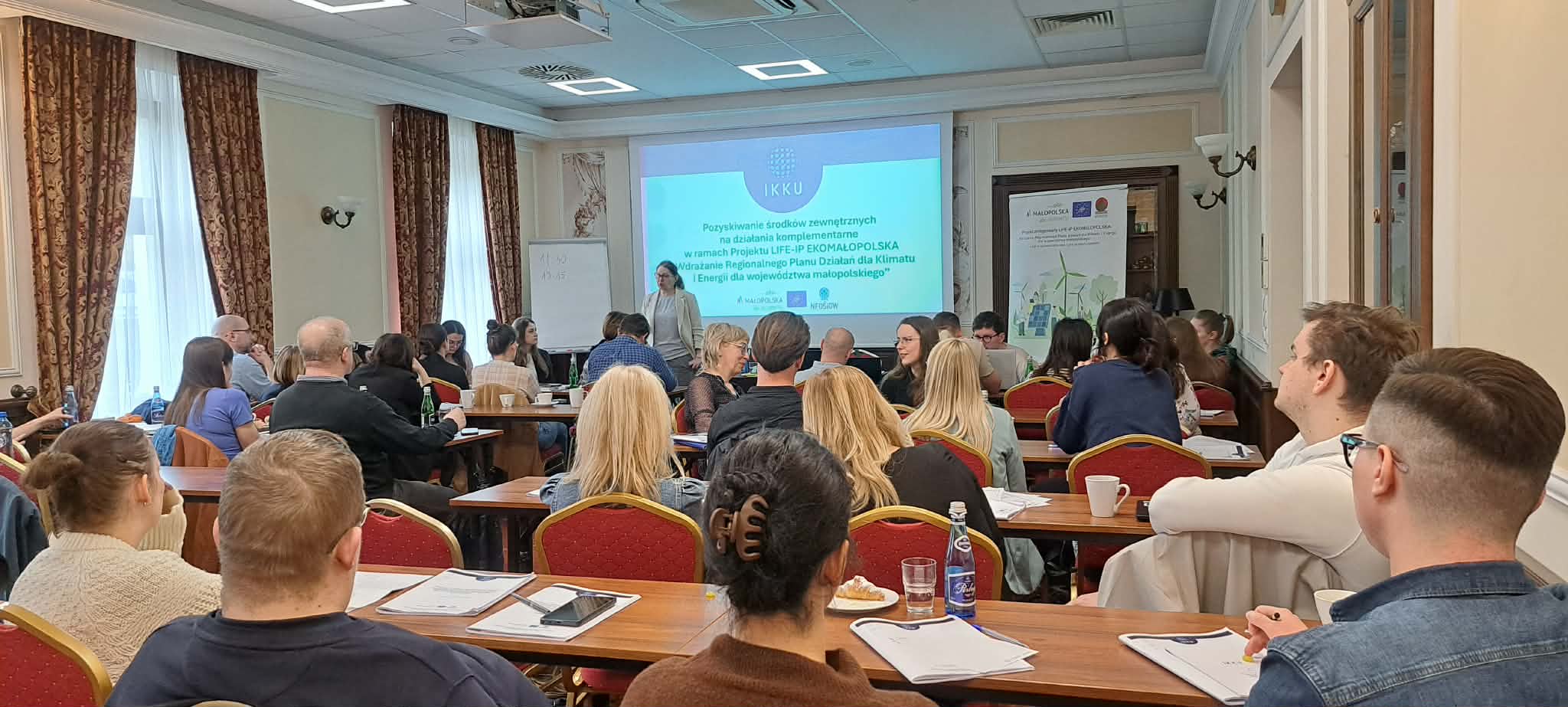 Duża grupa ludzi w ławkach ukazana od tyłu słucha wykładu ekspertki na drugim planie zdjęcia