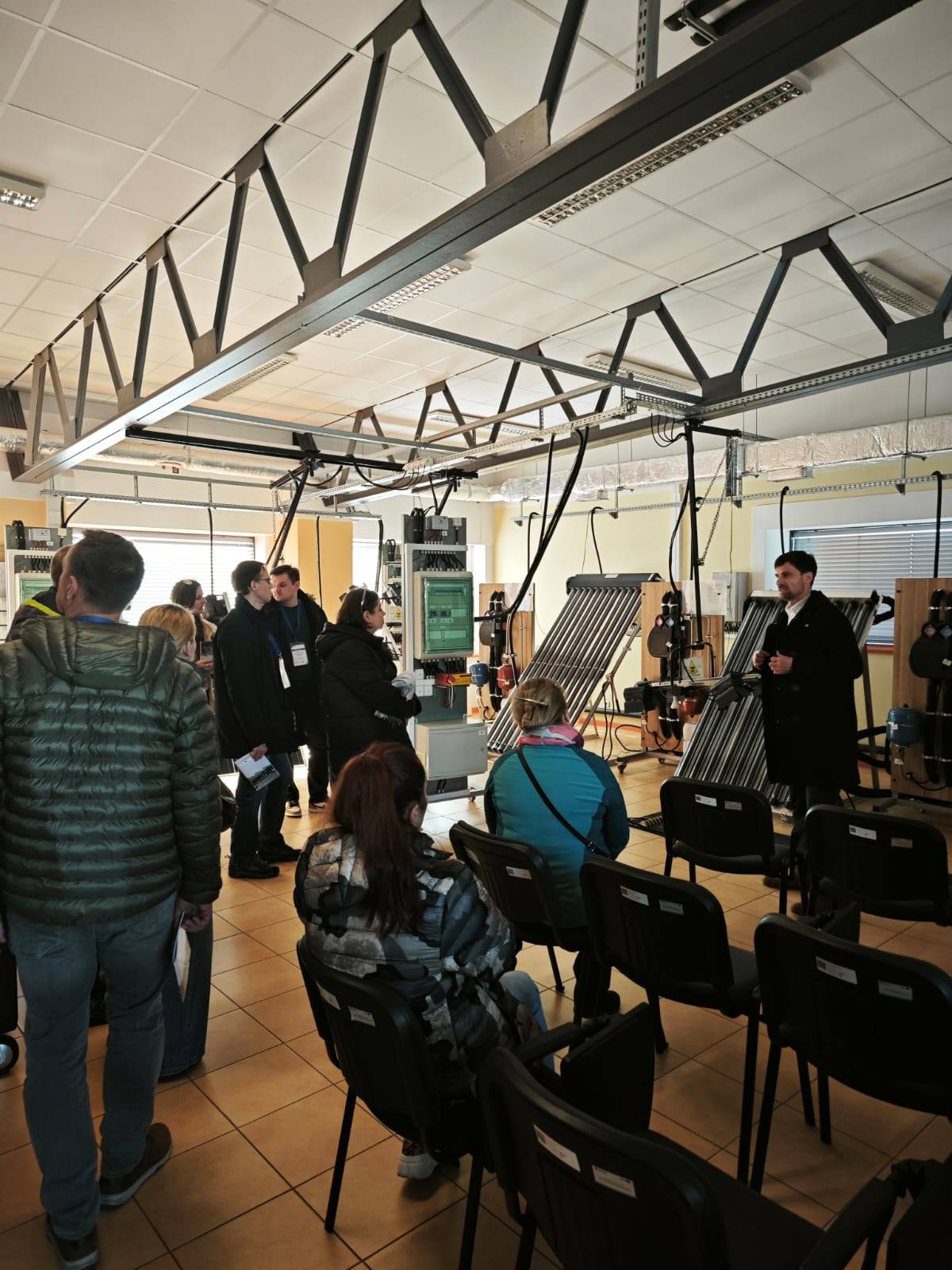 Grupa ludzi stoi lub siedzi na sali, w której prowadzony jest wykład nt. instalacji OZE.