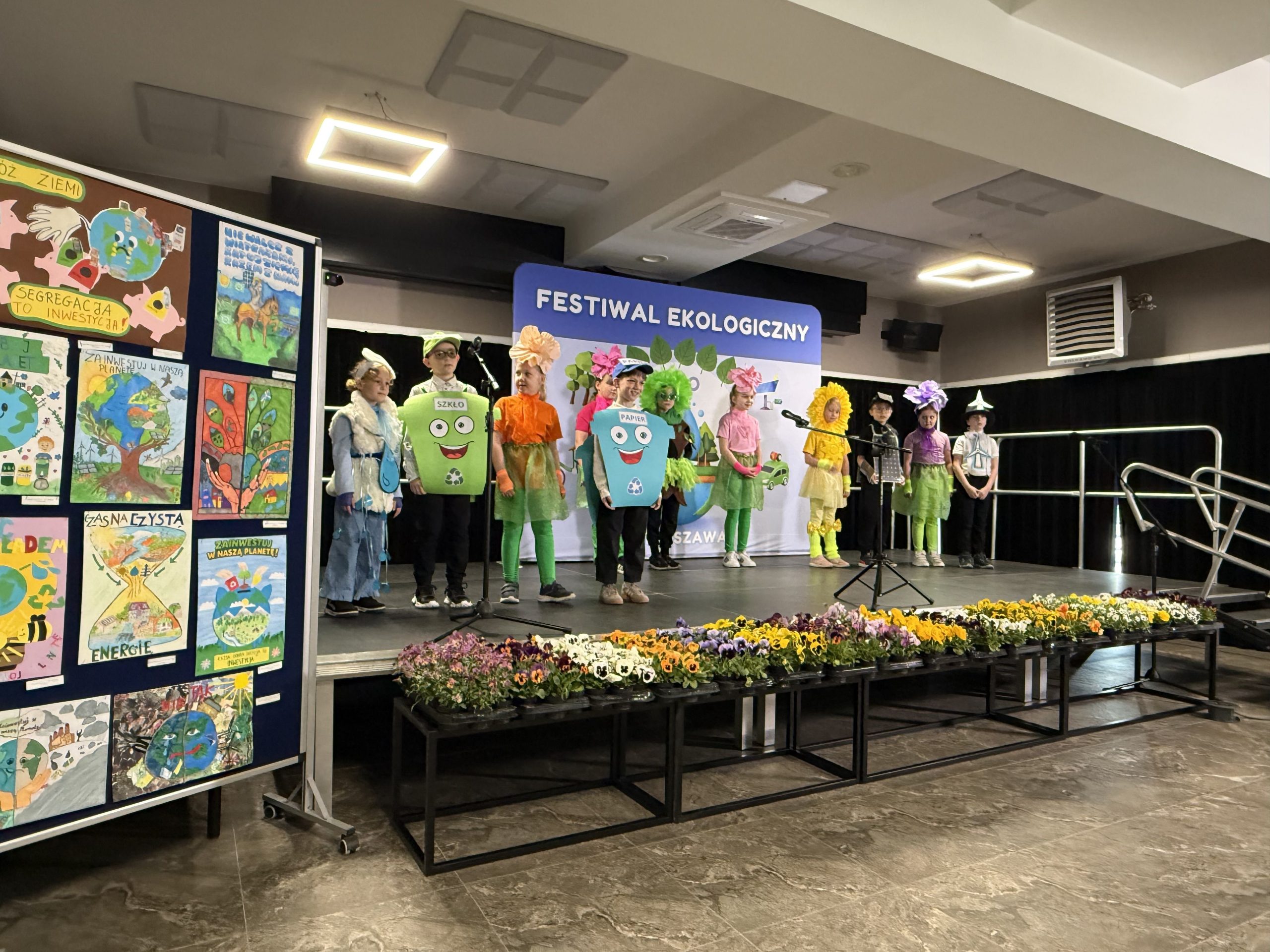 Grupa dzieci przebranych za kolorowe kosze i kwiaty stoi w rzędzie śpiewając piosenkę