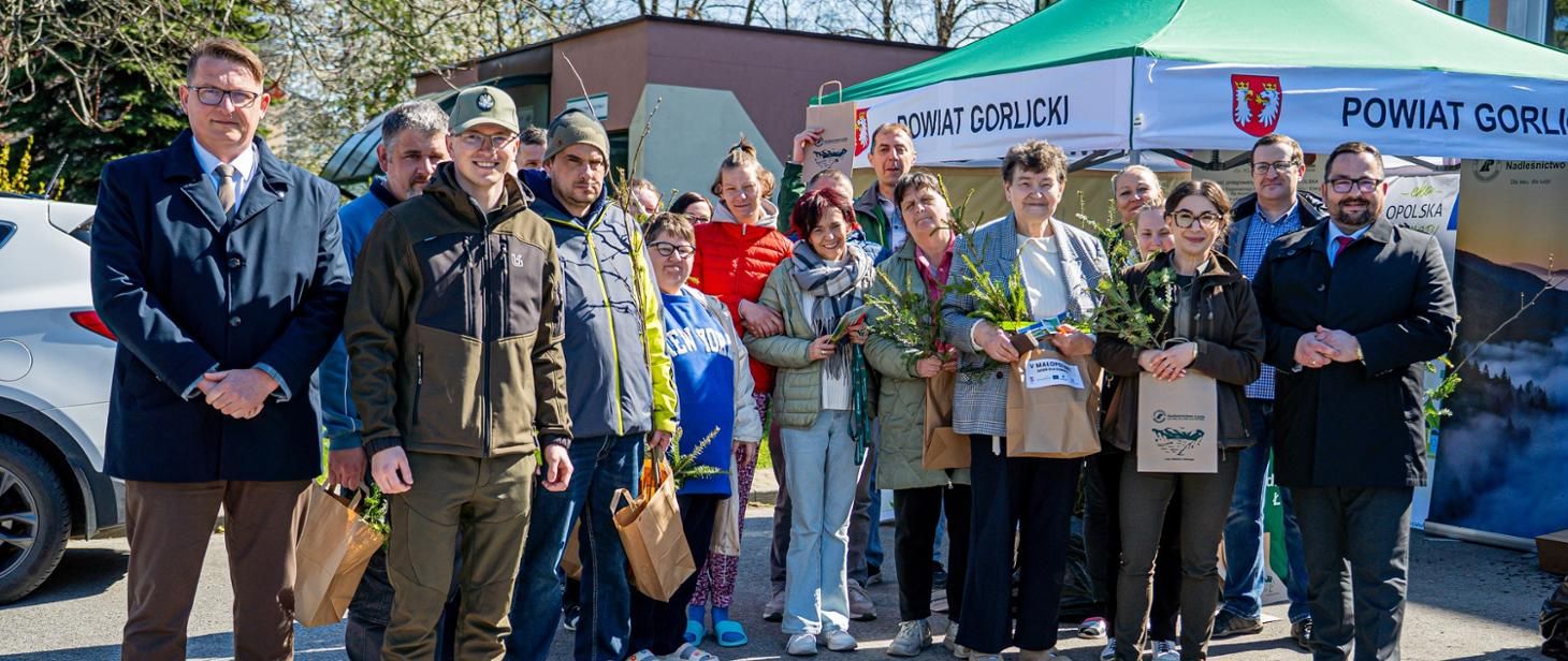 Grupa ludzi w różnym wieku pozuje do zdjęcia trzymając w papierowych torbach przekazane im sadzonki drzew iglastych 