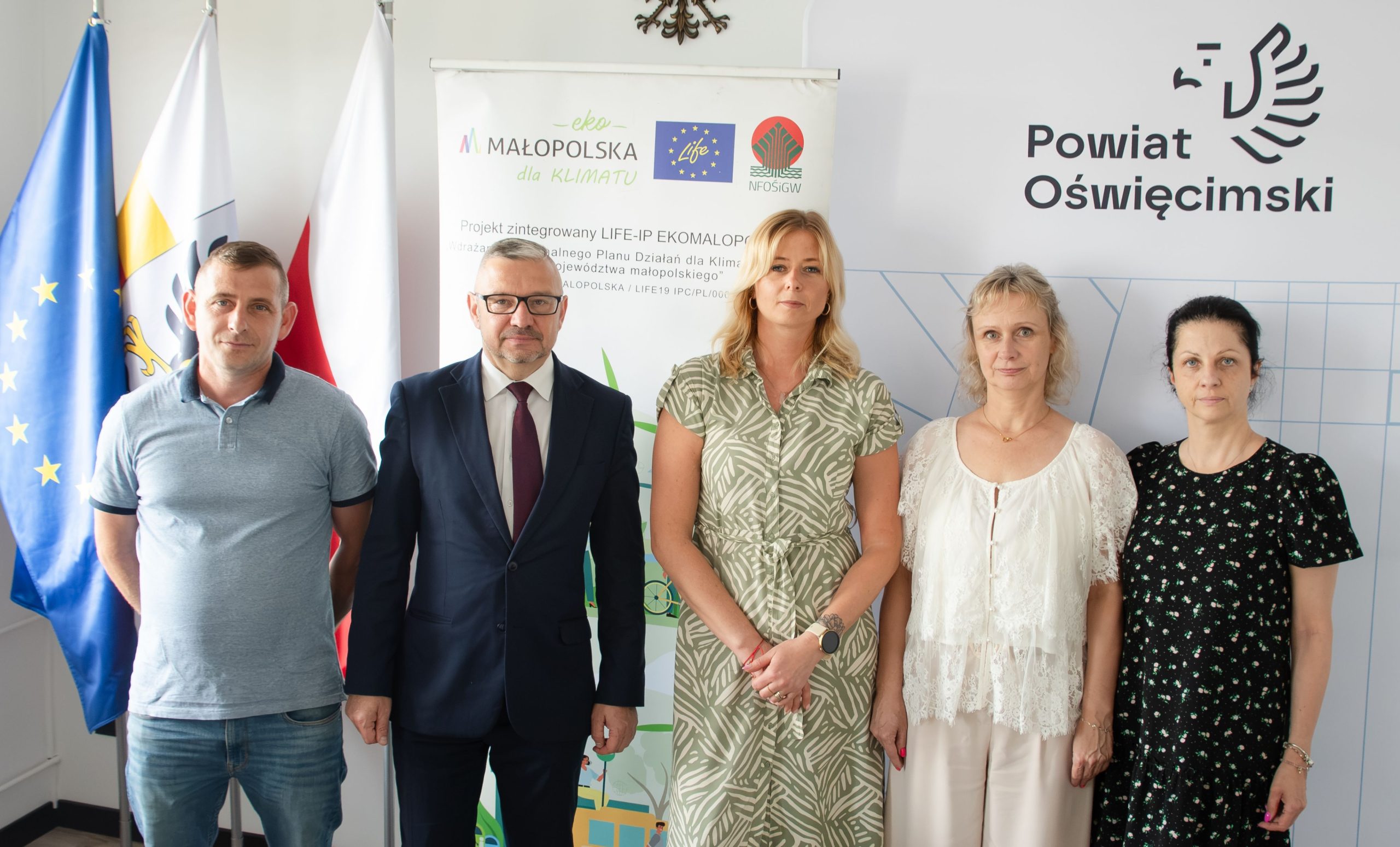 grupa ludzi stoi na tle roll-up'a z akronimem i grafika projektu, obok flaga polski, unii europejskiej i powiatu oświęcimskiego